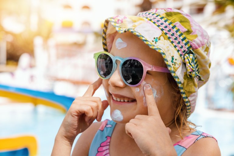 A young girl in a coloful hat and rainbow sunglasses smiling as she applies white sunblock on her cheeks.
