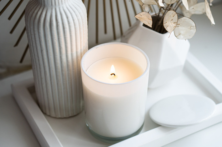 A nice white candle burning on a decorative dish next to other white decorations, such as a vase and florals.
