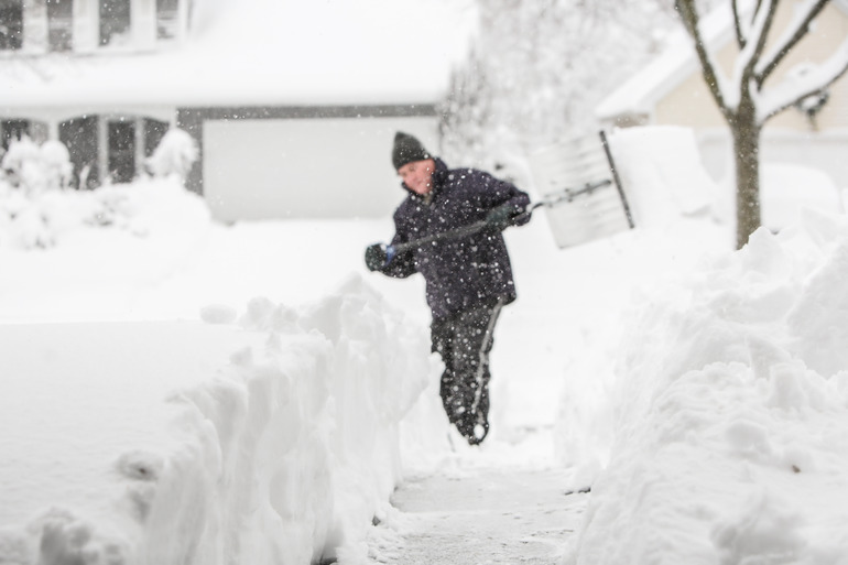 A man shoveling snow after a heavy snowfall. It's snowing as he shovels and he's in a black snowsuit and beanie.