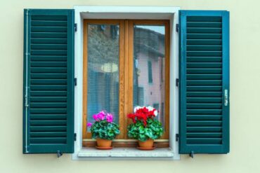 Closeup of windows on a home’s exterior with bright teal shutters and pink, white, and red potted flowers on the windowsill.