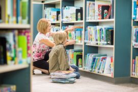There is a woman touching books on a bookshelf. There is also a child sitting next to her with a stack of books nearby.
