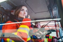A female wearing a red and yellow firefighter uniform is sitting in the passenger seat of a fire truck and holding a radio.