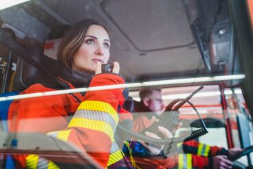 A female wearing a red and yellow firefighter uniform is sitting in the passenger seat of a fire truck and holding a radio.