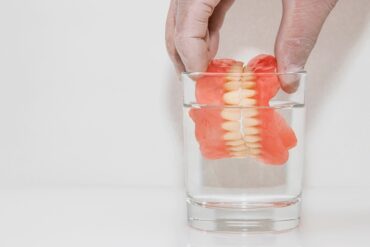 An individual is submerging top and bottom dentures in a clear glass of water on a white background.