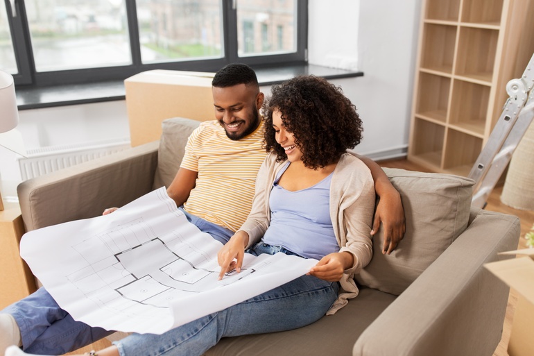 A happy couple sitting on a couch together, smiling, while they look at an extensive blueprint of a house floorplan.