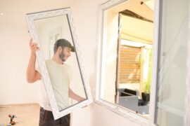 A young man wearing a black baseball cap holds a large white window while standing in front of an empty window frame.