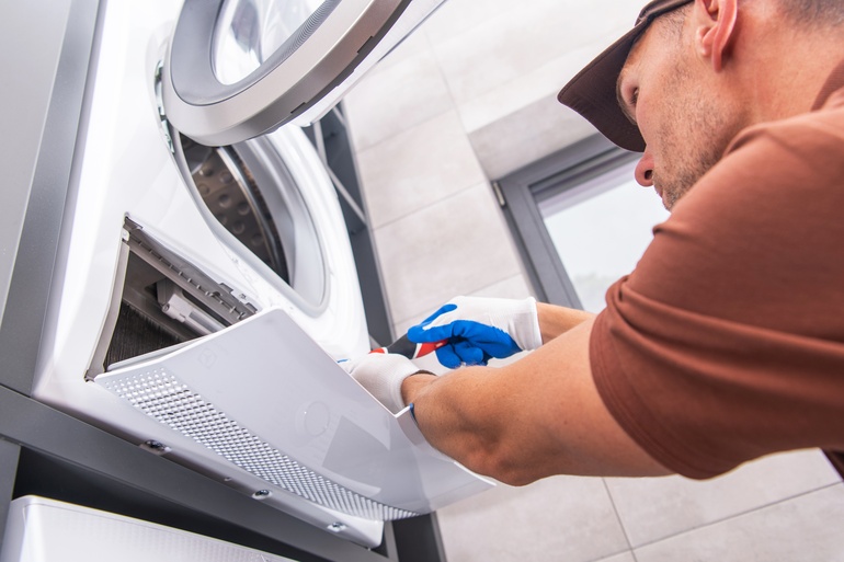 A man works on his broken washing machine with a screwdriver while wearing white and blue gloves and a baseball cap.