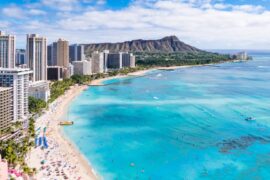 A view of Waikiki Beach in Oahu on a sunny, partly cloudy day. The water is bright blue, and beachgoers dot the shore and surf.