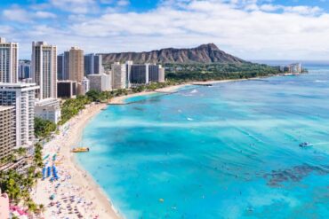 A view of Waikiki Beach in Oahu on a sunny, partly cloudy day. The water is bright blue, and beachgoers dot the shore and surf.