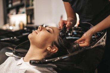 A woman leans back in a chair with her head in a black hair-washing salon bowl. A person is washing her hair.