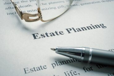 A close-up of the words "Estate Planning" on a piece of paper with glasses and a ballpoint pen lying on top.