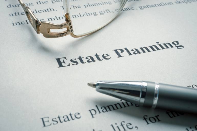 A close-up of the words "Estate Planning" on a piece of paper with glasses and a ballpoint pen lying on top.