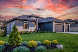 The exterior of a dark-colored house with a freshly cut lawn and lush landscaping scattered throughout the yard.