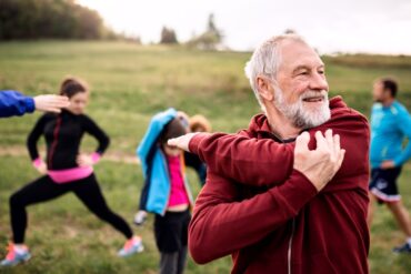 A group of people are in a park and stretching with an older man in front stretching his arm and shoulder.