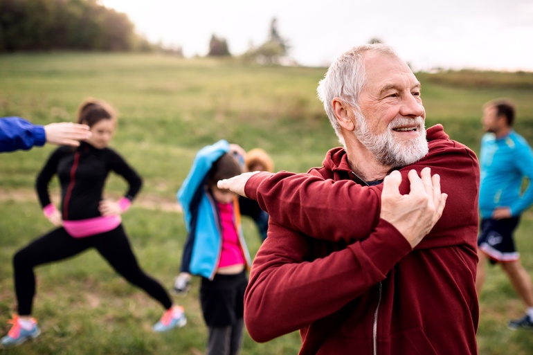 A group of people are in a park and stretching with an older man in front stretching his arm and shoulder.