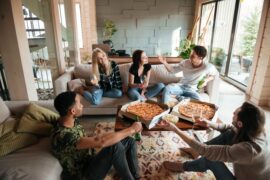 A group of friends are sitting in a living room, laughing. Some are holding drinks, and there are pizzas on the coffee table.