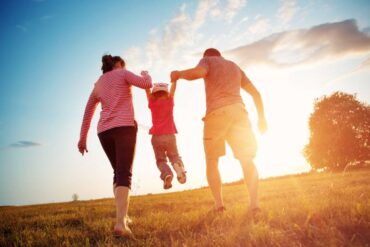 A small child with their parents. One parent is holding each arm on the child and swinging them in the air as the sun sets.