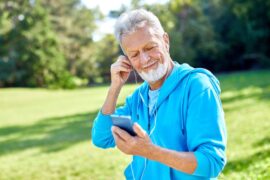 A smiling senior man places an earbud in his ear while holding his smartphone. He stands outside at a park on a sunny day.