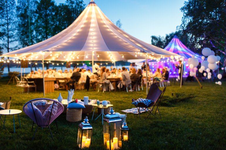An intimate seating area with comfortable chairs, pillows, and lanterns appears outside near an event tent with people.