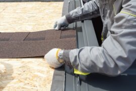 Construction worker wearing grey uniform and safety gloves holding an asphalt tile sheet on the roof of a home.