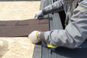 Construction worker wearing grey uniform and safety gloves holding an asphalt tile sheet on the roof of a home.