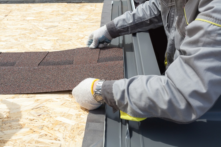 Construction worker wearing grey uniform and safety gloves holding an asphalt tile sheet on the roof of a home.