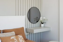 A gray floating shelf appears next to a bed with brown bedding. The shelf contains a white pot with white flowers.