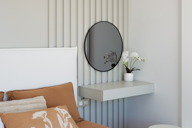 A gray floating shelf appears next to a bed with brown bedding. The shelf contains a white pot with white flowers.