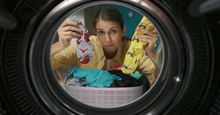 A confused woman holding up two mismatched patterned socks while looking into the drum of a washer.