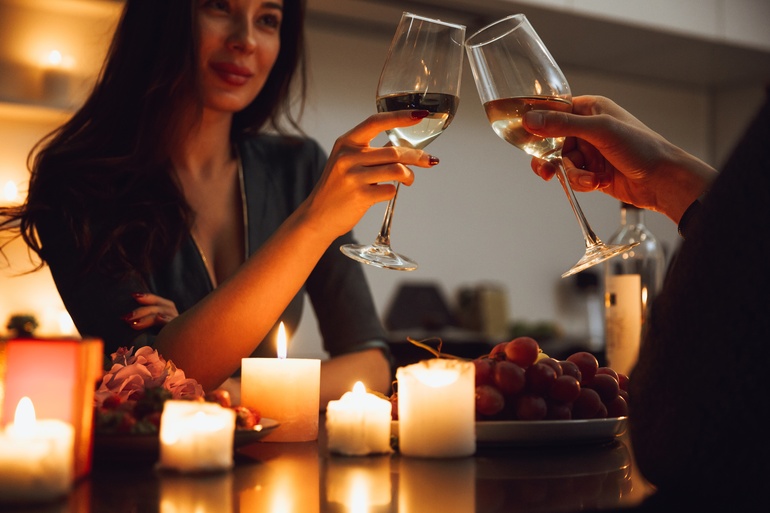A woman sits at a table across from another person, clinking two glasses of wine. There are candles and grapes on the table.