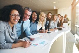At a long table in an office, multiple employees lean against the table, all smiling toward the camera.