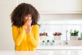 A woman wearing a yellow sweater stands in her kitchen and has a disgusted look on her face, holding her nose shut.