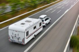 A white car with a roof carrier pulls a white RV down a highway. The road and surroundings are blurred by movement.