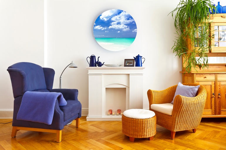A living room with two chairs and a faux fireplace. A circular metal canvas with a photo of a beach on it hangs on the wall.