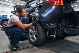A man wearing a gray shirt and blue overalls using a tool on a dark-colored vehicle's tire well during a repair.