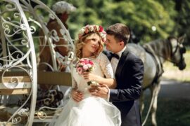 A man in a black tuxedo wraps his arms around a woman in a wedding dress from behind. They stand next to a horse and carriage.