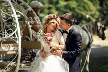 A man in a black tuxedo wraps his arms around a woman in a wedding dress from behind. They stand next to a horse and carriage.