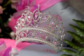 A beautiful silver tiara with crystals in it sits on a pink and white surface with the word “Mis” below the crown.