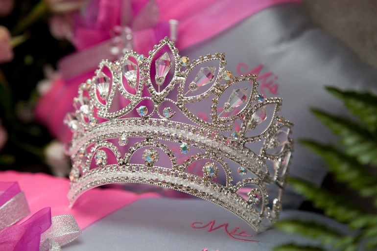 A beautiful silver tiara with crystals in it sits on a pink and white surface with the word “Mis” below the crown.