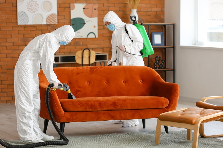 Two people wearing white biohazard suits and blue masks are cleaning a house. One uses a vacuum, and the other uses a sprayer.