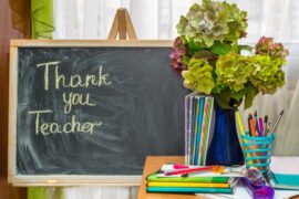 The words "Thank you, Teacher" written on a chalkboard, next to a desk with school supplies and a vase of hydrangeas on it.