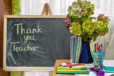 The words "Thank you, Teacher" written on a chalkboard, next to a desk with school supplies and a vase of hydrangeas on it.