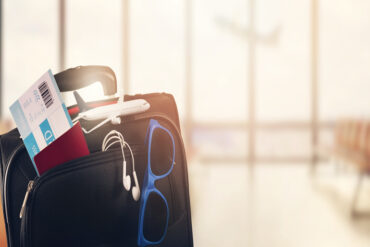 A close-up view of a black luggage with a miniature airplane toy, wired earbuds, plane ticket, and a blue sunglasses.