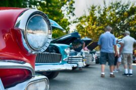 A close-up view shows the headlight of a red, old vehicle parked next to other vintage cars. People are walking.