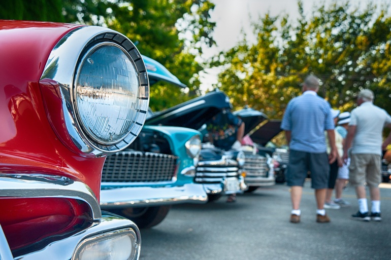 A close-up view shows the headlight of a red, old vehicle parked next to other vintage cars. People are walking.
