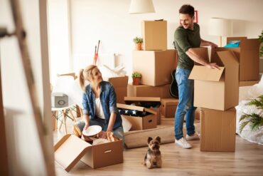 A man and woman pack stacks of cardboard boxes in a well-lit living room. A small dog sits between them.