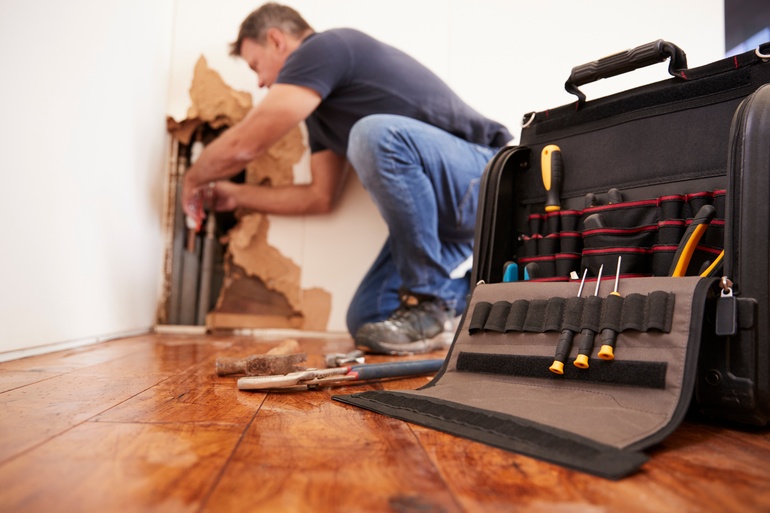 A worker is kneeling one knee while they work on pipes in the corner of a room, with their tool bag in front of them.