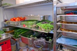 The inside of a walk-in freezer. The shelves are loaded with metal trays full of fresh produce and finished dishes.