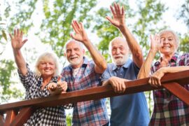 Four older people smiling and waving from behind a wooden railing. There are green trees behind them.