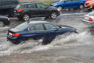 On a rainy day, several cars on the road drive through intense water pouring down. One vehicle makes a small wave.